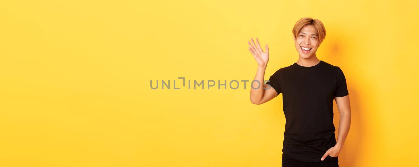 Portrait of handsome friendly asian guy in black outfit, waving hand to say hello and smiling, greeting someone, standing yellow background by Benzoix