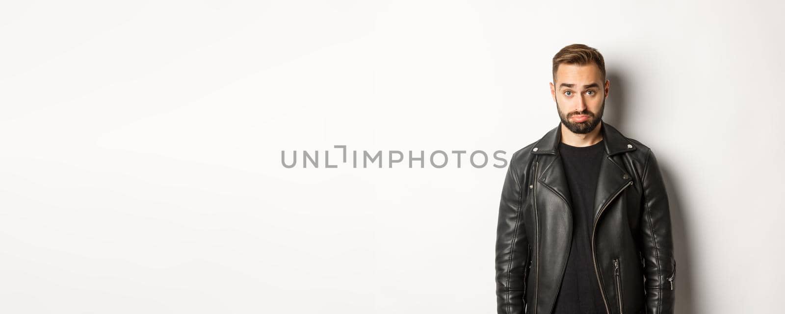 Sad and cute guy in leather black jacket pouting, looking silly at camera, standing against white background.