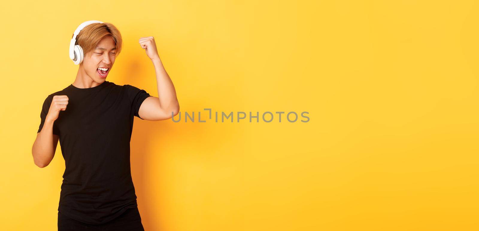 Carefree attractive asian guy with blond hair, singing along and dancing as listening to music in wireless headphones, standing yellow background by Benzoix