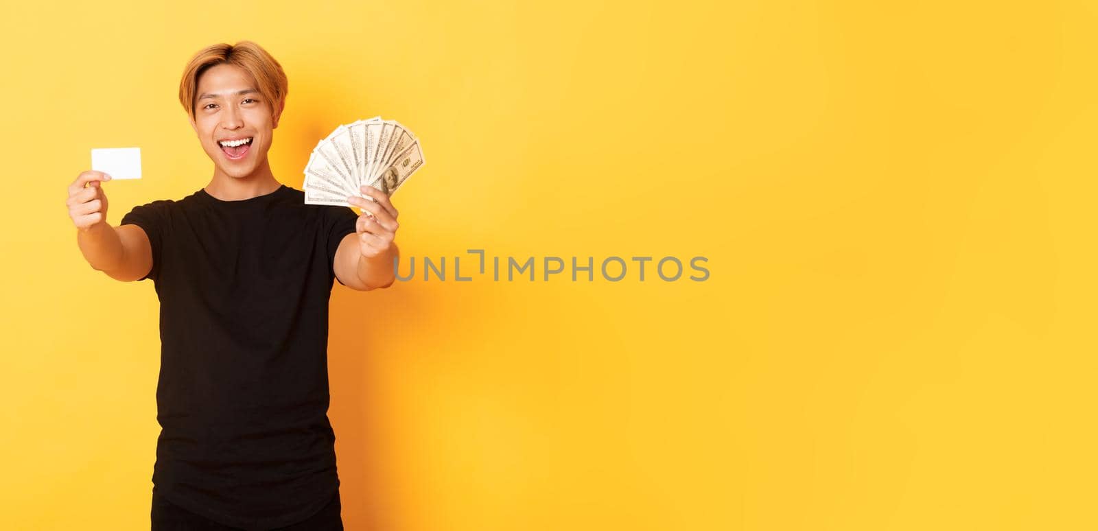 Happy good-looking asian guy in black casual clothes, showing money and credit card, smiling sassy, yellow background by Benzoix