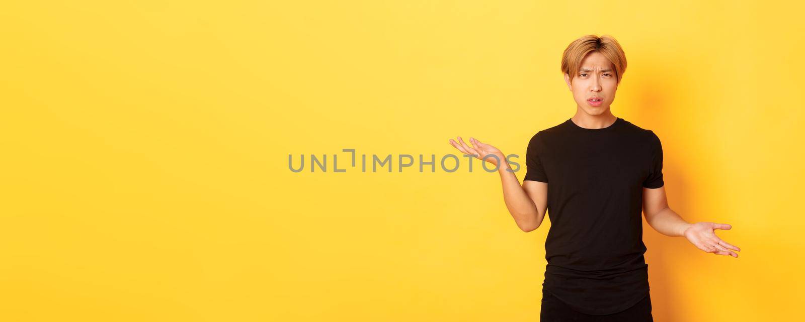 Portrait of confused and annoyed asian guy spread hands sideways, cant understand something, standing yellow background by Benzoix