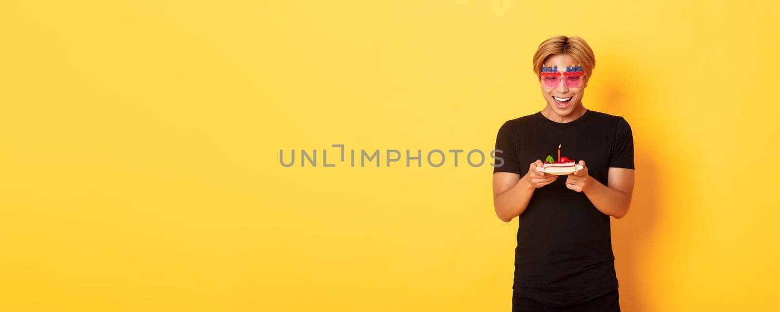 Excited handsome asian guy in party glasses, looking hopeful at birthday cake as celebrating b-day, making wish on lit candle, standing yellow background.