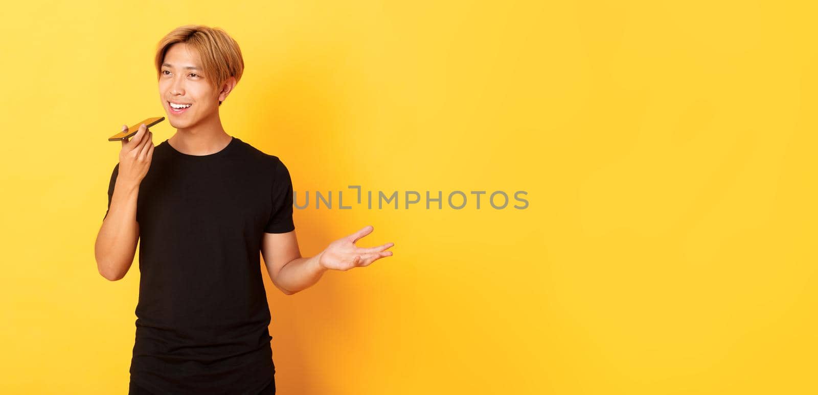 Portrait of stylish handsome korean guy with blond hair record voice message on mobile phone, holding smartphone near mouth and speaking, standing yellow background.