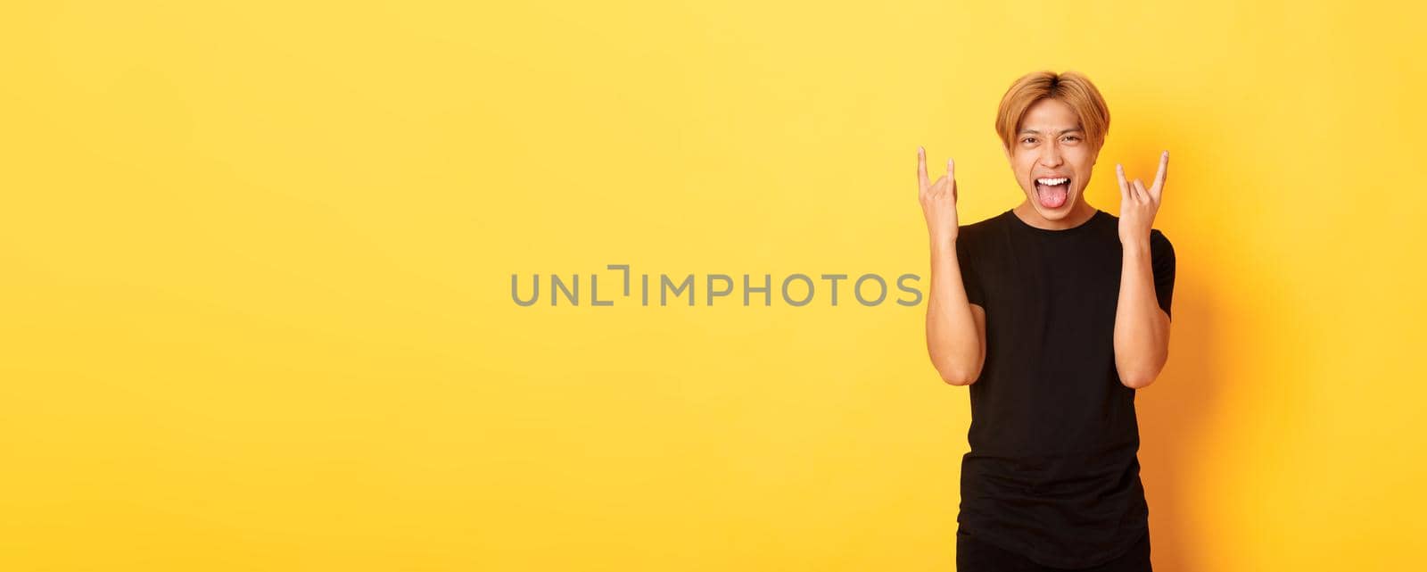 Portrait of joyful handsome asian blond guy having fun, showing rock-n-roll gesture and stick tongue carefree, standing yellow background.