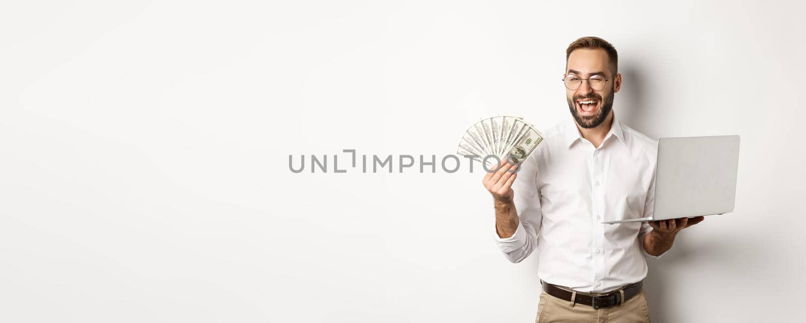 Business and e-commerce. Confident businessman showing how work online, winking, holding money and laptop, standing over white background.