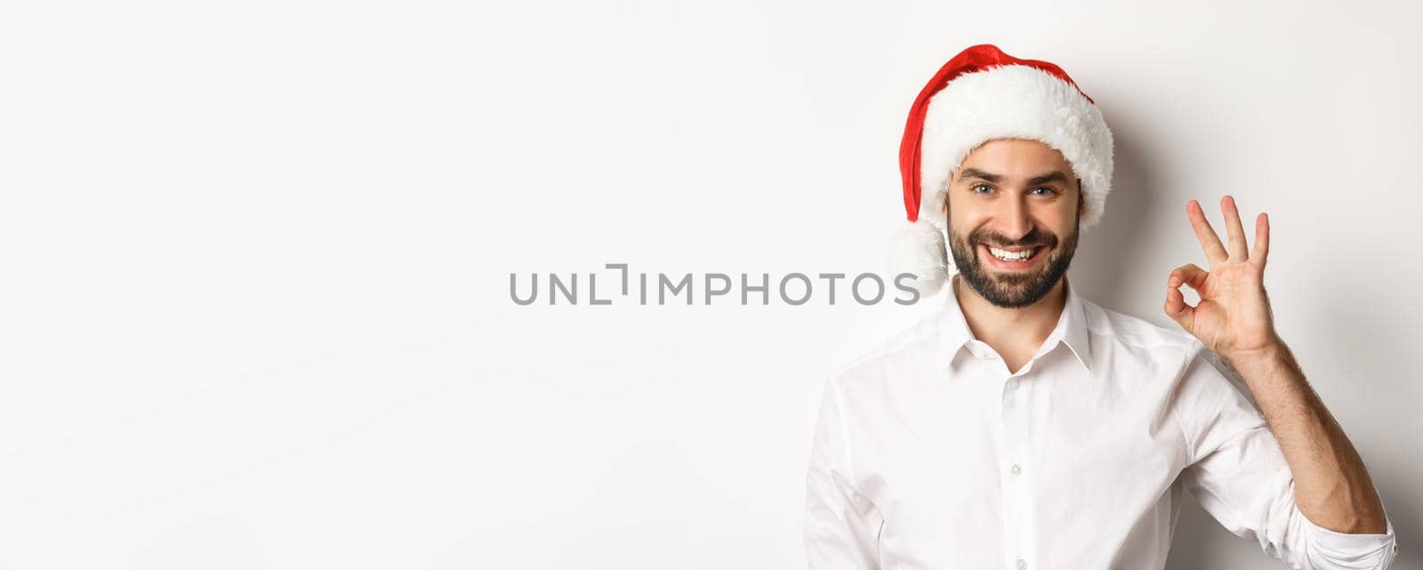 Party, winter holidays and celebration concept. Confident man in santa hat showing okay sign, approve and like, white background.