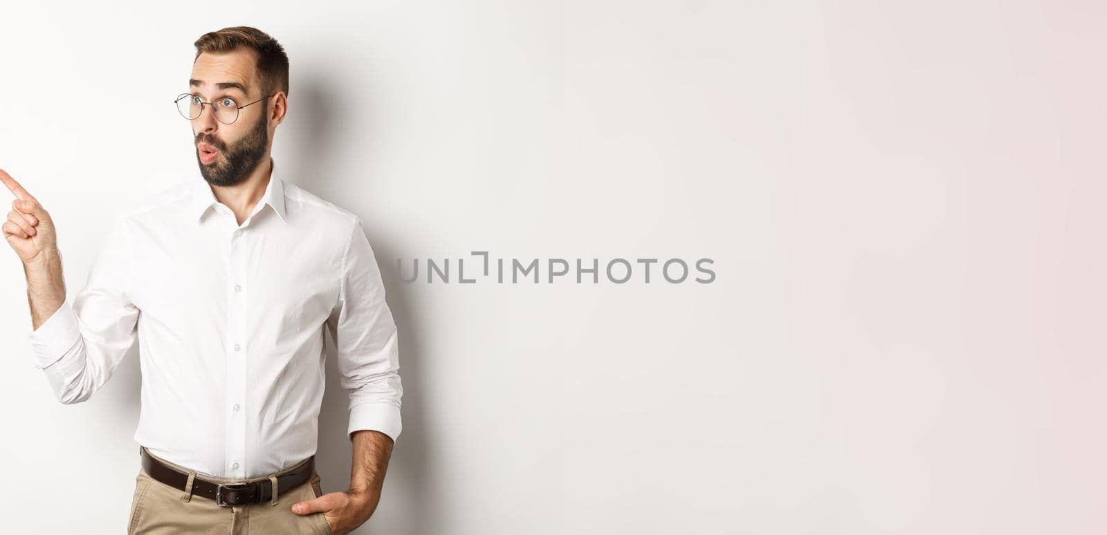 Intrigued young man in glasses pointing finger left, looking at promo offer interested, standing against white background.