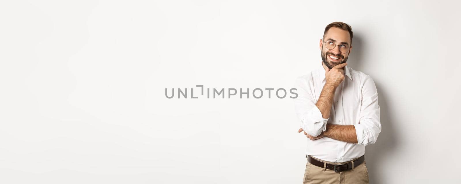 Satisfied businessman having interesting idea, looking at upper left corner and thinking with pleased smile, standing over white background.