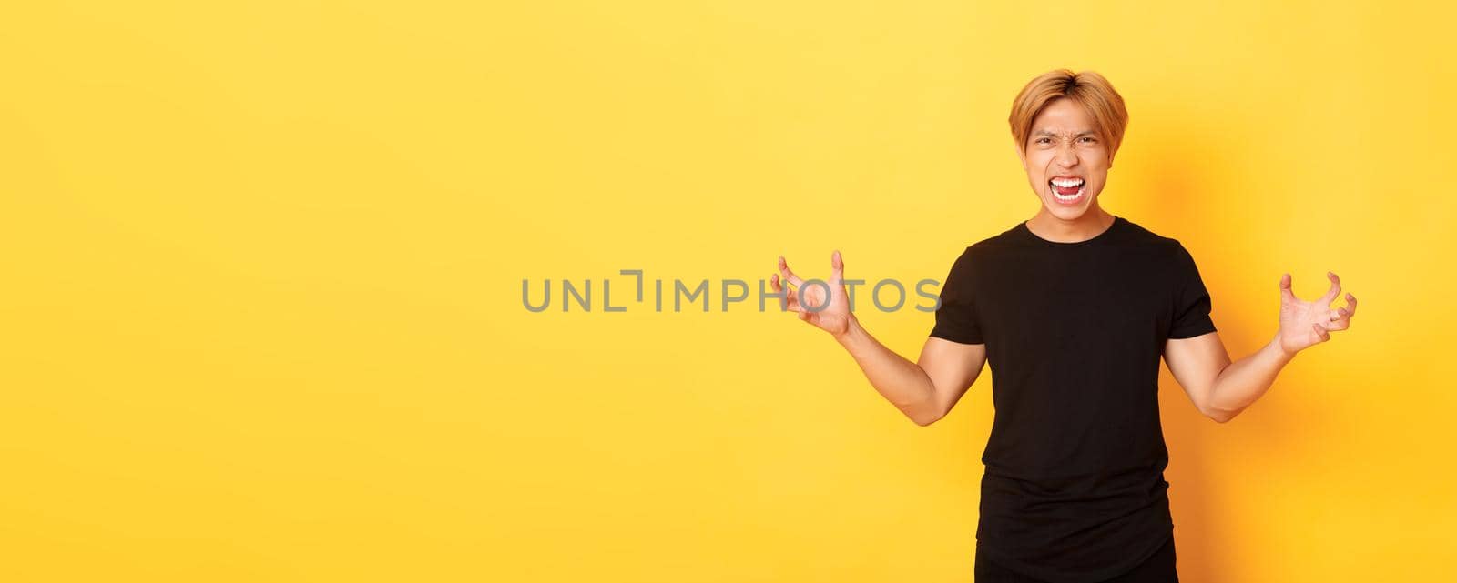 Portrait of outraged, pissed-off asian man shaking hands hateful and grimacing, shouting angry, standing over yellow background.