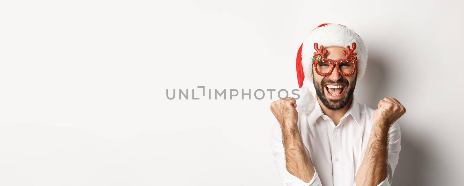 Close-up of man celebrating christmas or new year, wearing xmas party glasses and santa hat, rejoicing and shouting of joy, standing over white background.