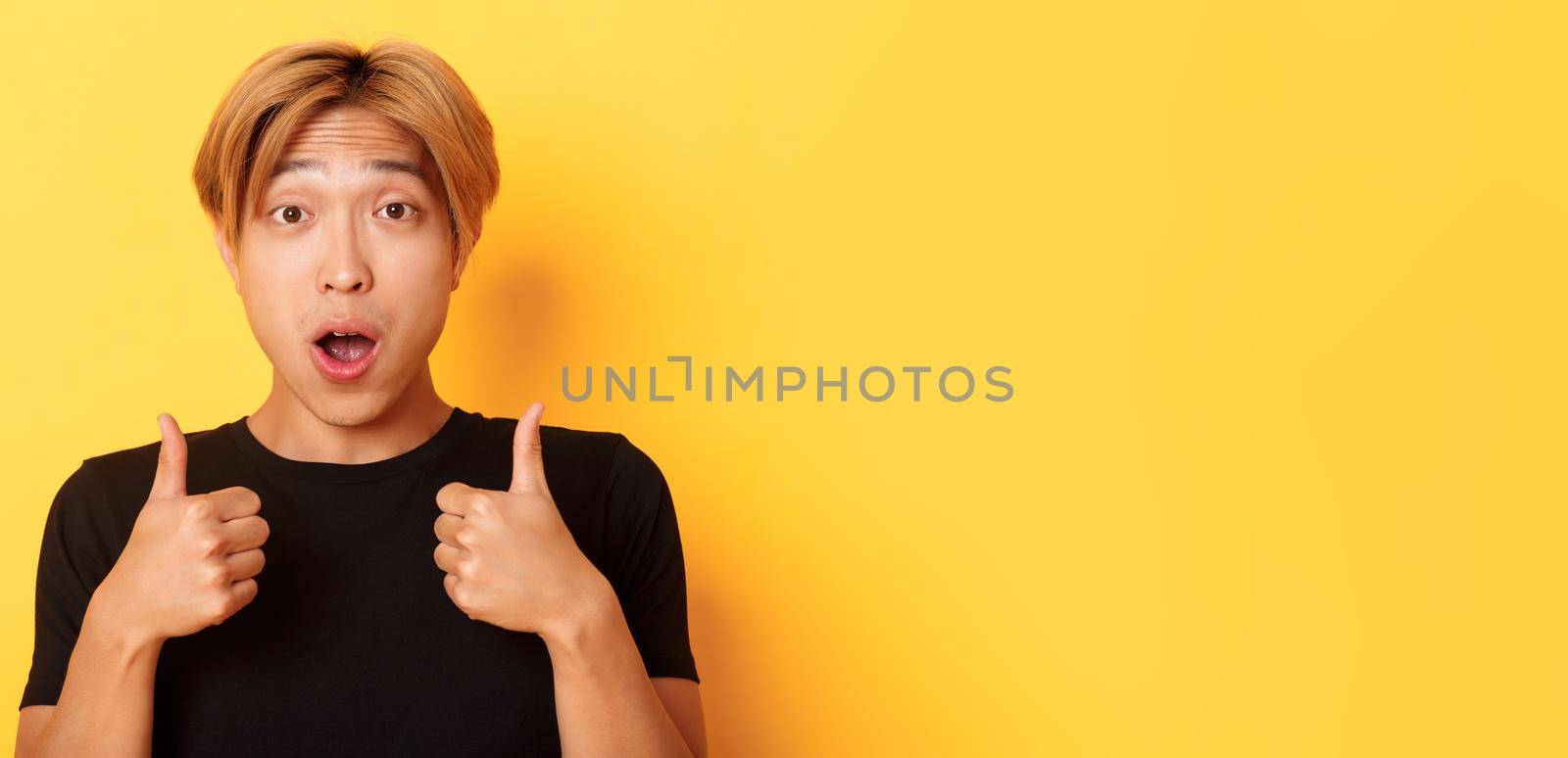 Close-up of impressed and excited handsome asian guy showing thumbs-up and looking amazed, standing over yellow background.