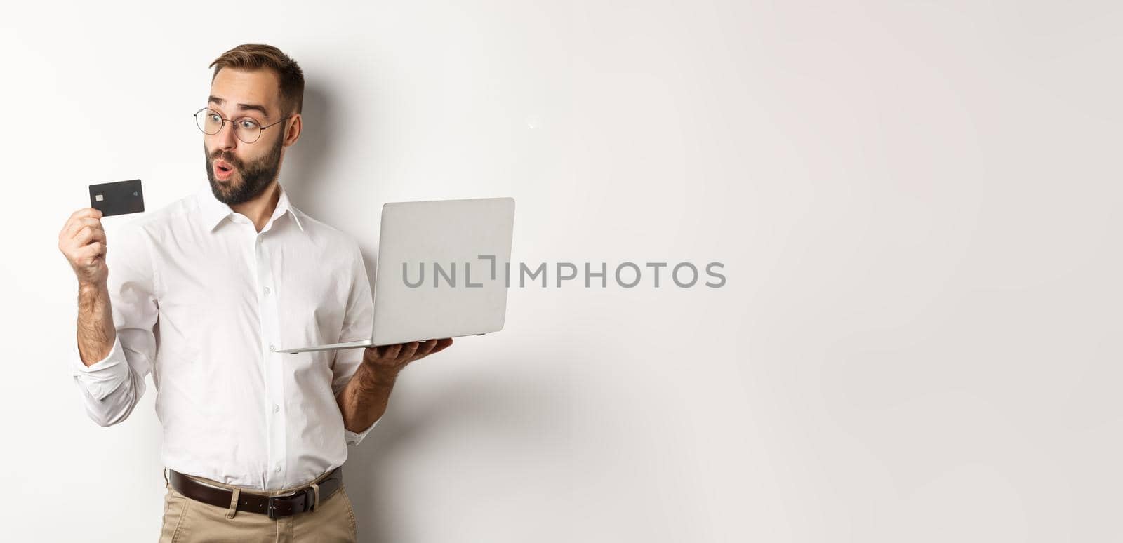 Online shopping. Amazed businessman holding laptop, looking impressed at credit card, standing over white background by Benzoix