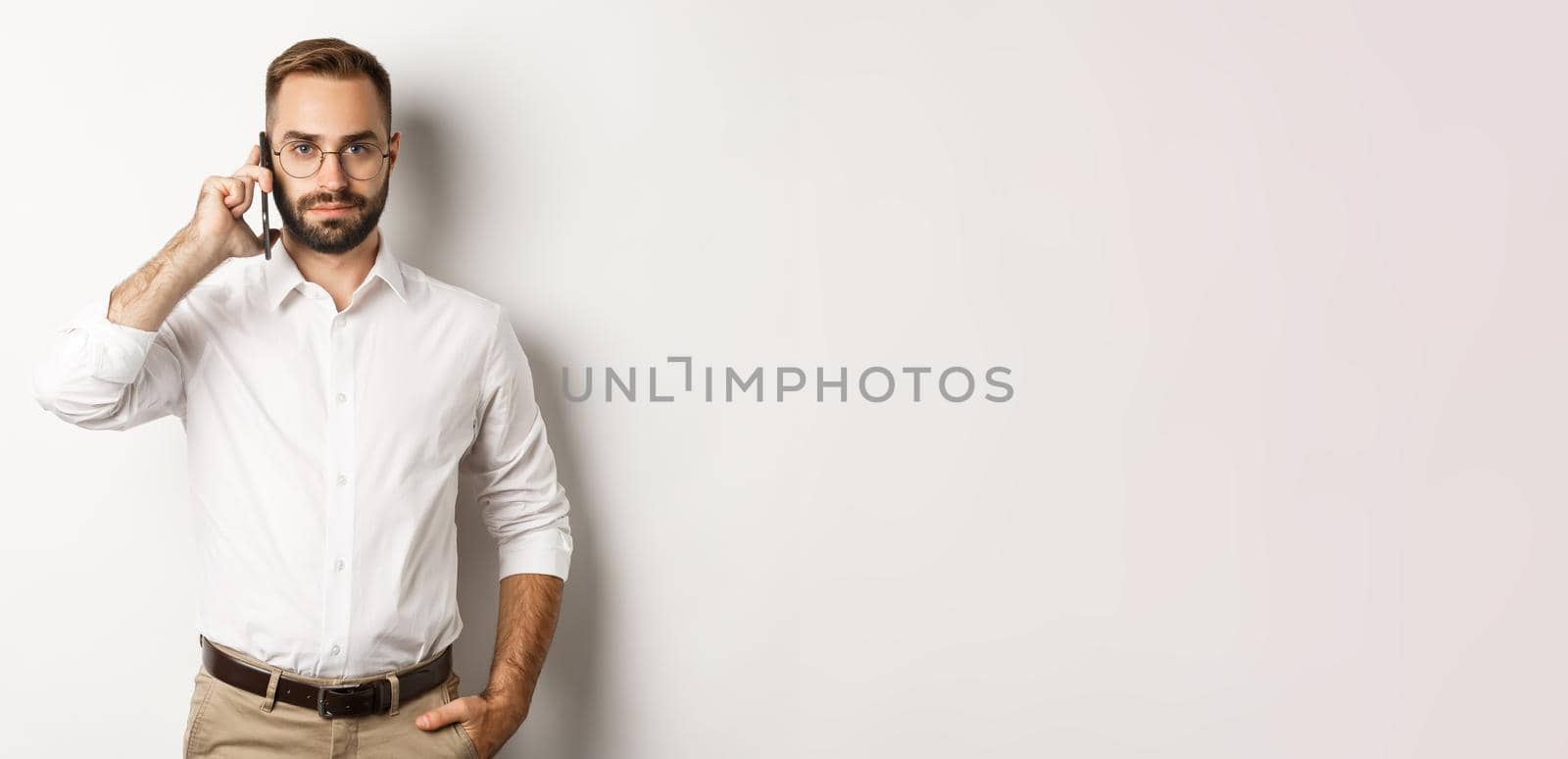 Confident business man talking on phone, looking serious, standing over white background.