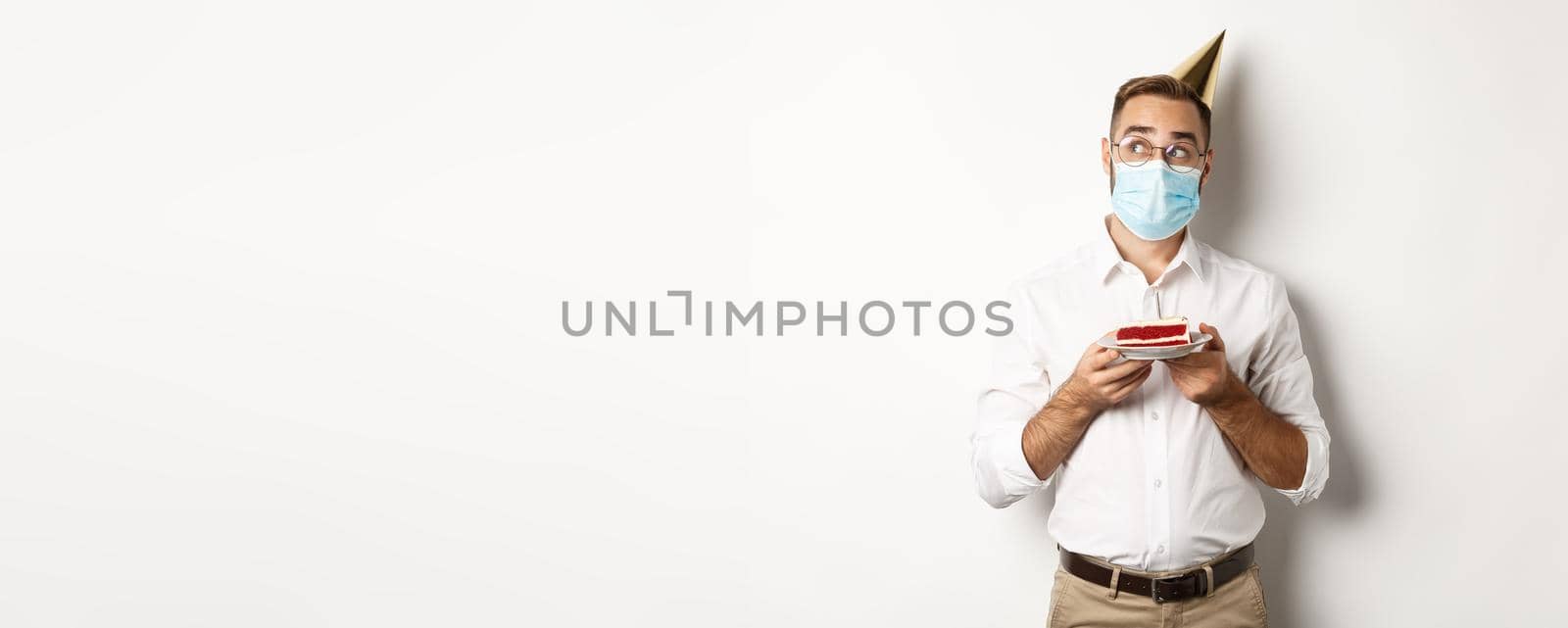 Covid-19, social distancing and celebration. Thoughtful man holding birthday cake, making wish and wearing face mask on quarantine, white background.