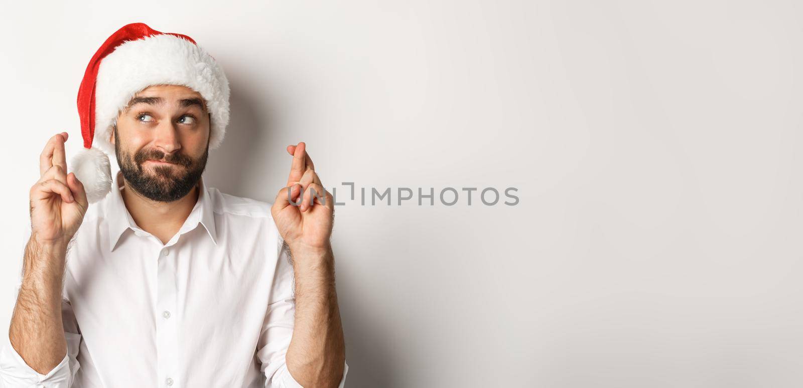 Party, winter holidays and celebration concept. Happy man in santa hat making christmas wish, cross fingers for good luck and looking hopeful at upper left corner.