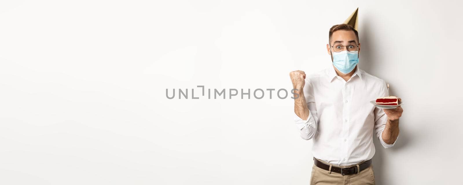 Covid-19, social distancing and celebration. Hopeful happy birthday man in face mask, holding bday cake and rejoicing, standing against white background.