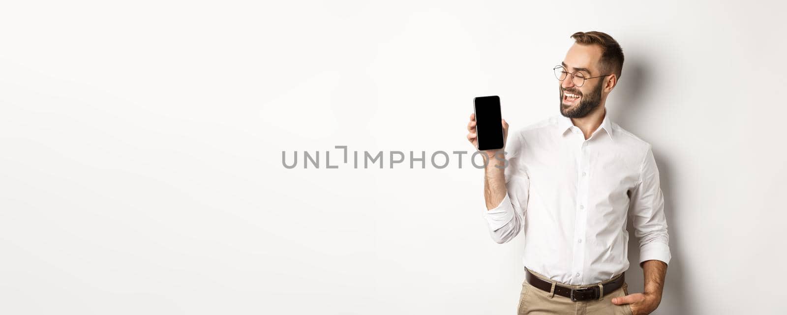 Satisfied business man showing and looking at mobile screen, introduce application or website promo, standing over white background.