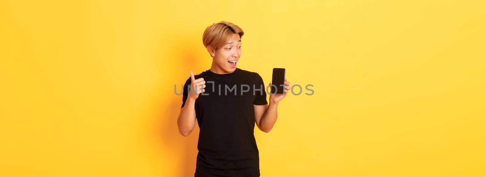 Portrait of satisfied asian guy looking at smartphone screen and showing thumbs-up in approval, standing yellow background by Benzoix