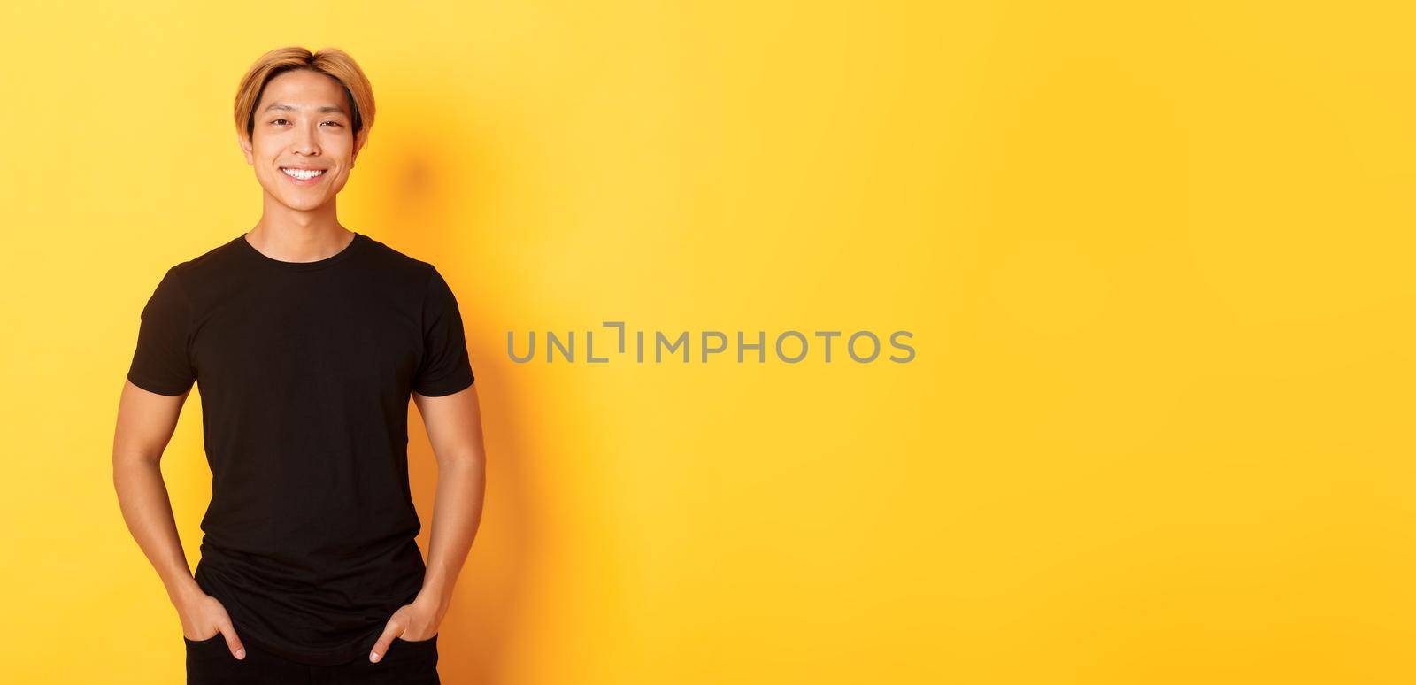 Attractive smiling asian man in black t-shirt, standing pleased, yellow background.