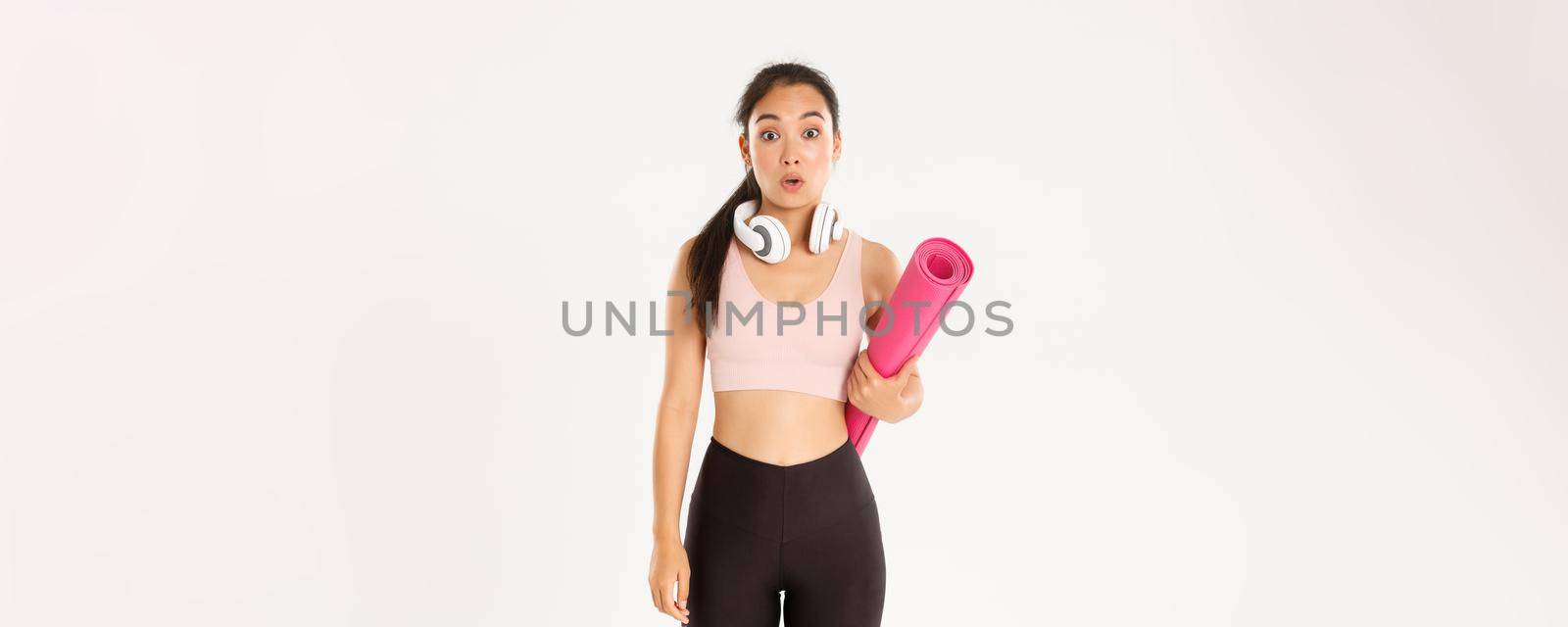 Sport, wellbeing and active lifestyle concept. Confused and shocked asian fitness girl, cant understand why gym closed, holding rubber mat and looking surprised at camera, white background.