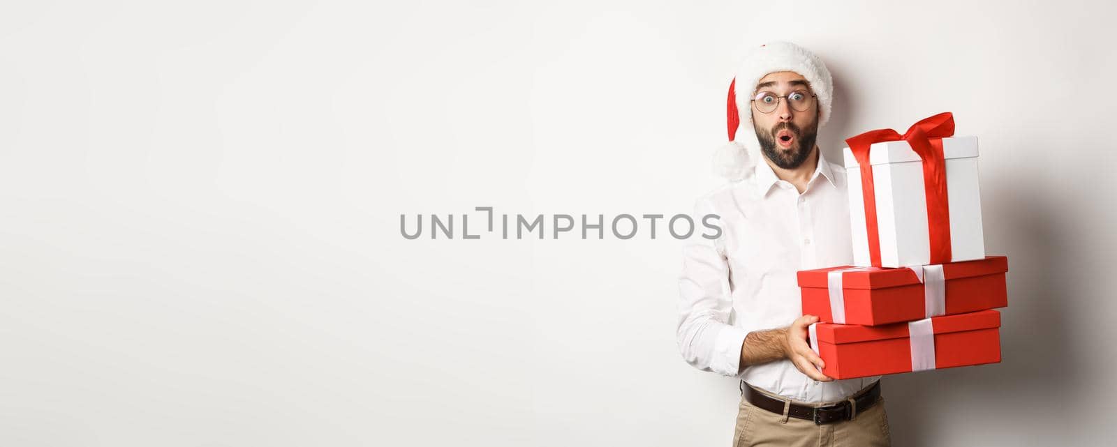 Winter holidays and celebration. Excited man holding Christmas gifts and looking surprised, wearing Santa hat, standing over white background.