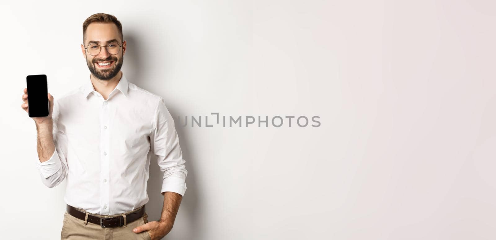 Satisfied business man showing mobile screen, smiling proudly, standing over white background.