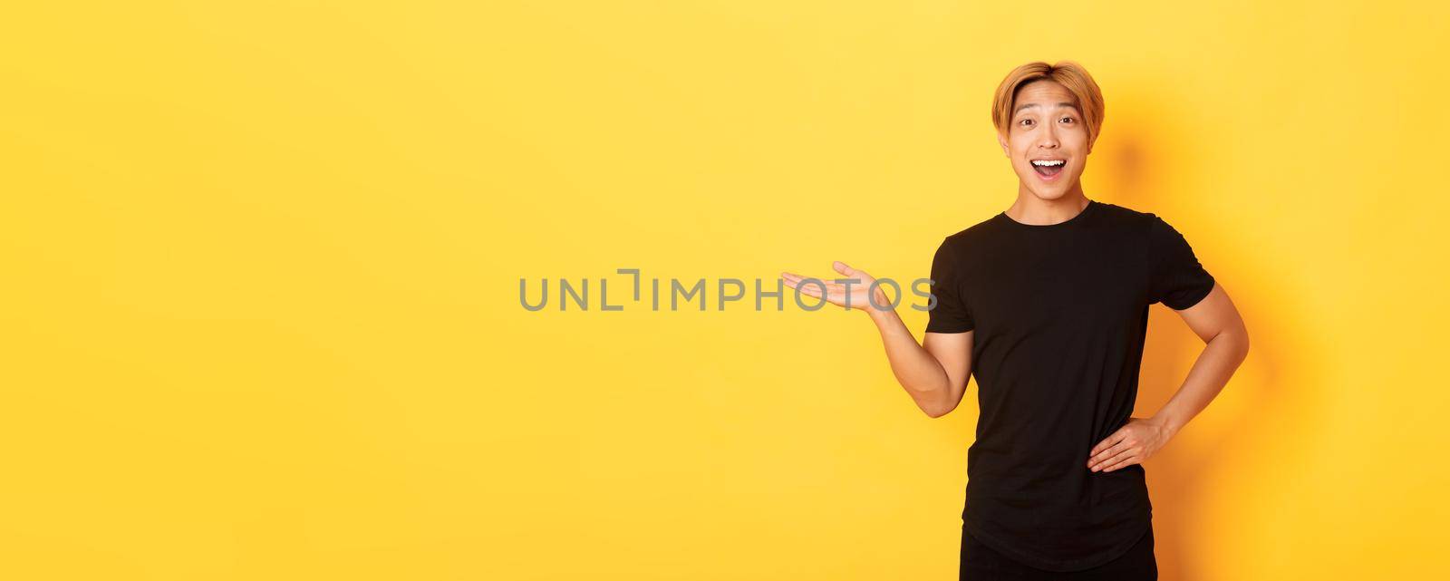 Portrait of happy and excited smiling asian guy holding something on hand over yellow background by Benzoix