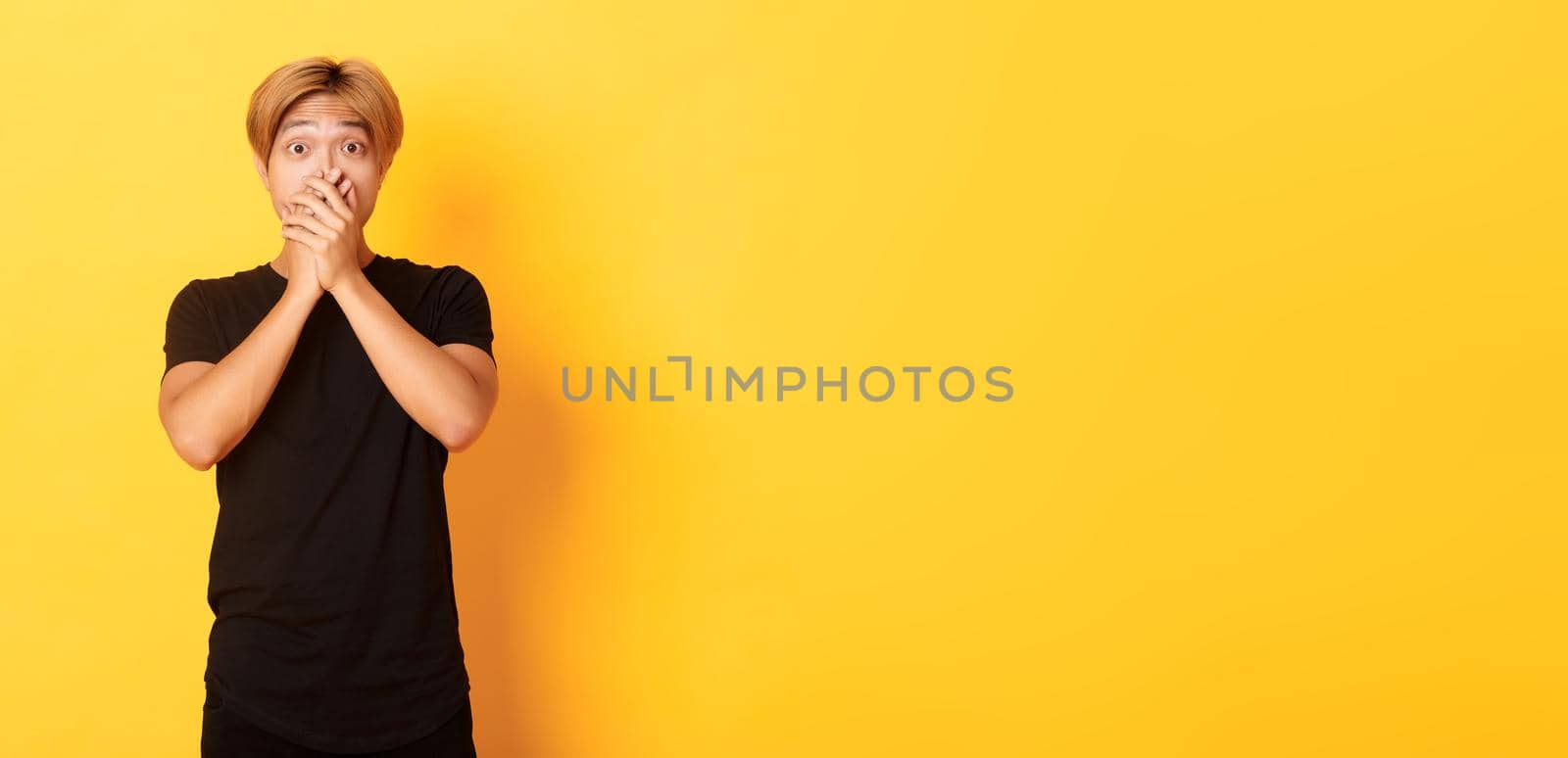 Portrait of surprised gasping asian guy with blond haircut, cover mouth with hands and looking amazed, standing yellow background by Benzoix