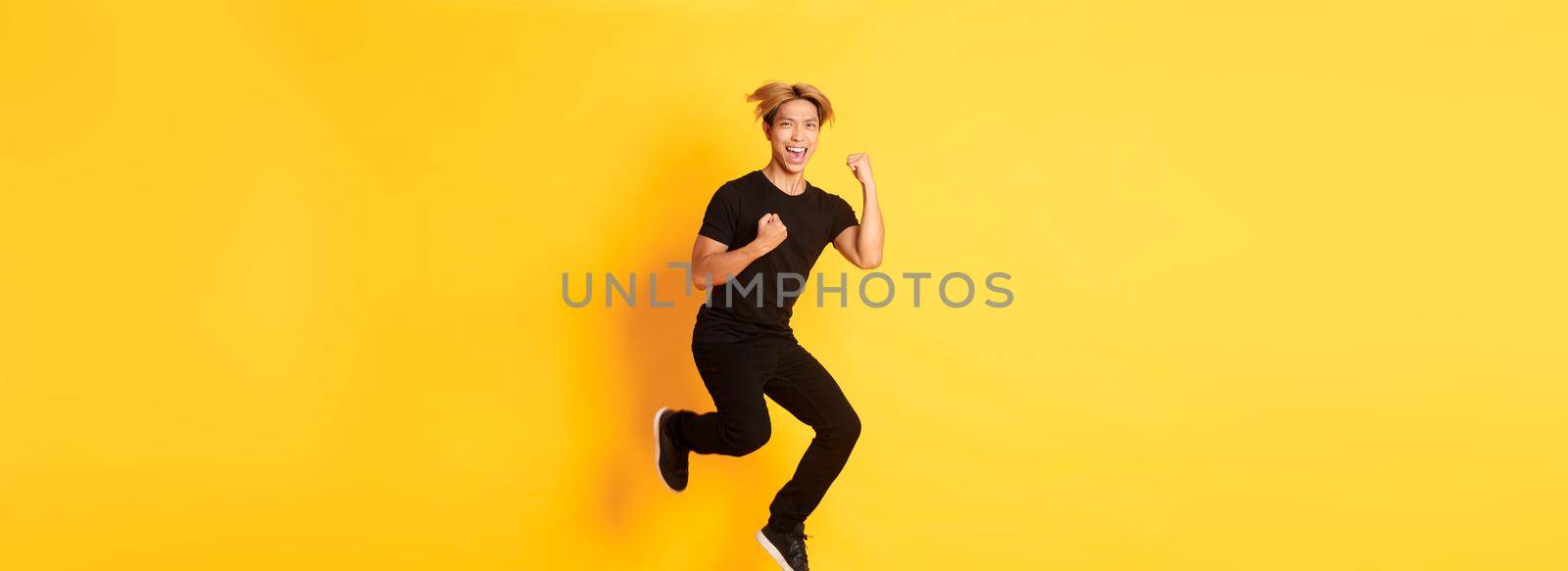 Full-length of happy attractive asian guy in black clothes jumping and celebrating victory, achieve goal, standing yellow background, triumphing.