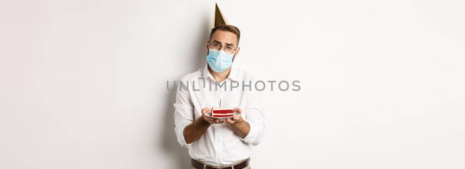 Coronavirus, quarantine and holidays. Sad man cant blow candle off birthday cake, wearing face mask and complaining, standing over white background by Benzoix