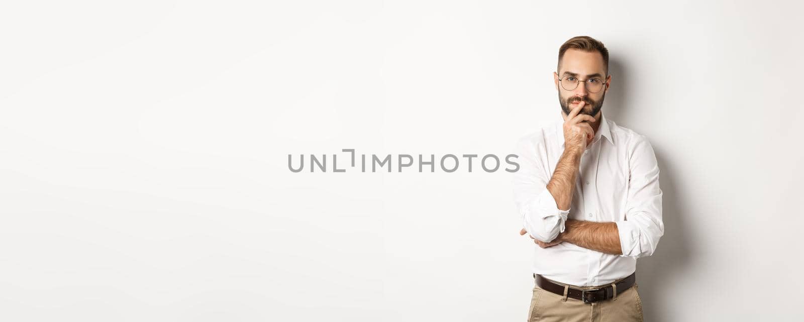 Thoughtful handsome businessman looking at camera, making choice or thinking, standing in glasses and white collar shirt against studio background.