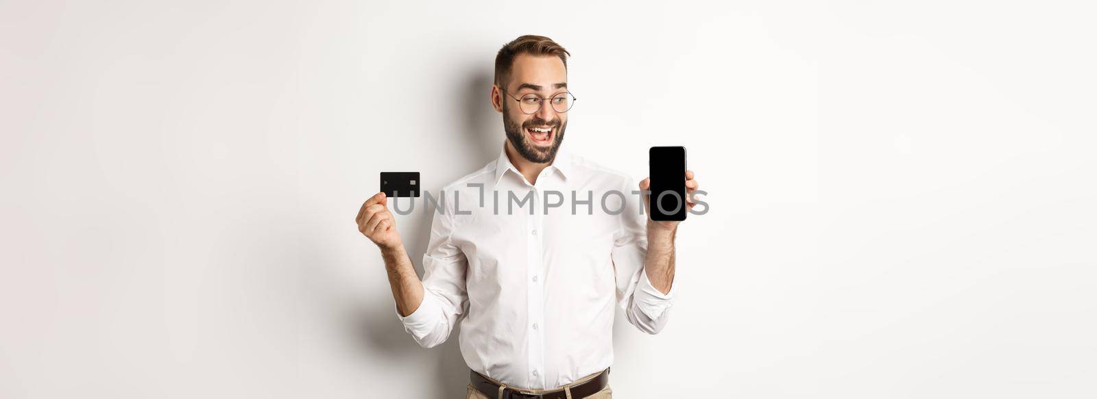 Business and online payment. Smiling handsome man showing mobile screen and credit card, standing over white background.