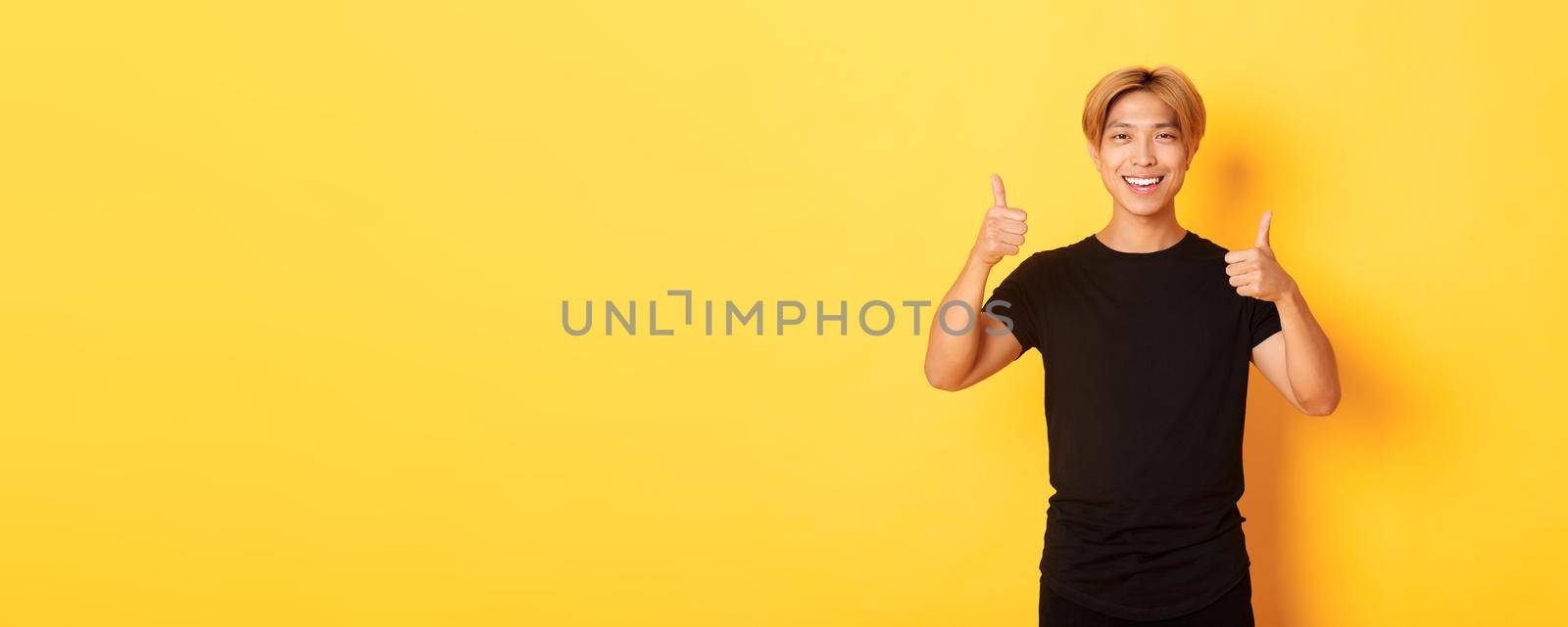 Portrait of satisfied asian man smiling and showing thumbs-up over yellow background.