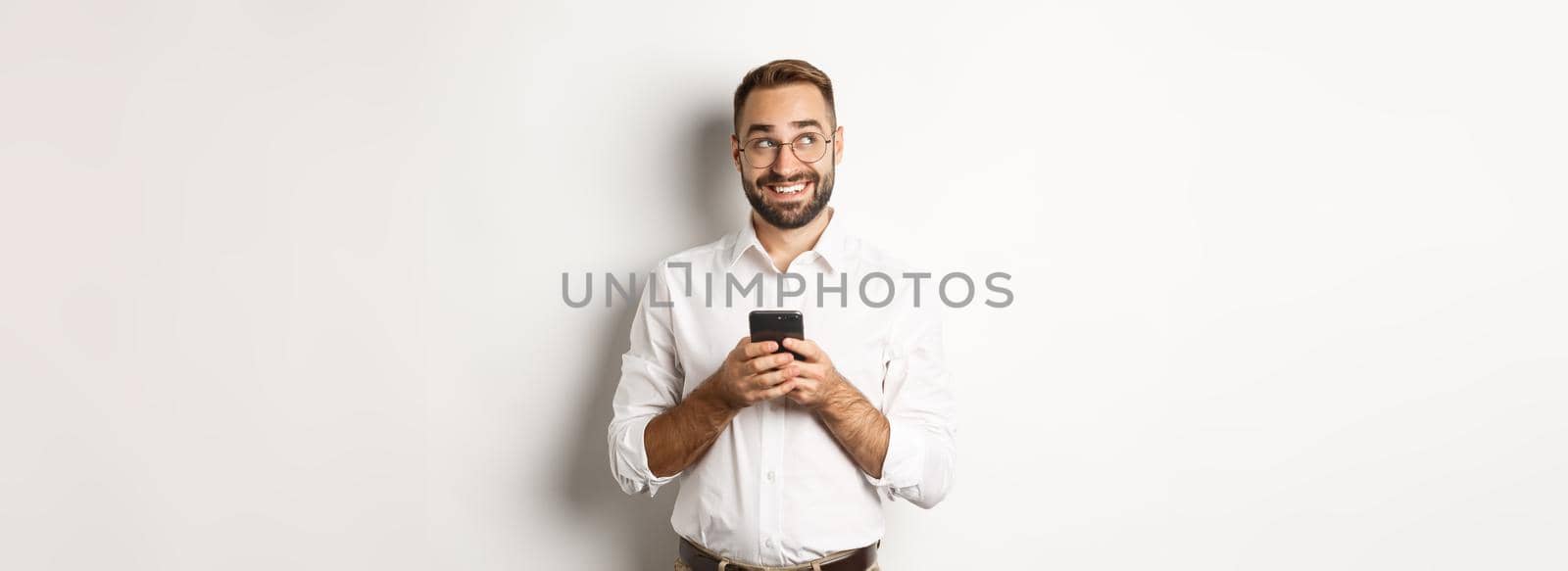 Thouthful handsome manager using mobile phone and thinking about answering message, looking at upper left corner and smiling, standing over white background.