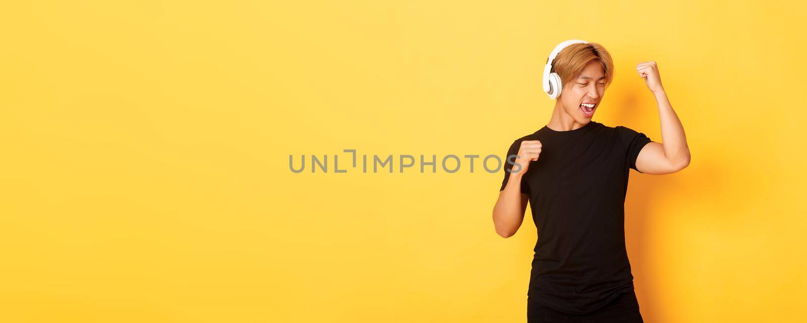 Carefree attractive asian guy with blond hair, singing along and dancing as listening to music in wireless headphones, standing yellow background by Benzoix