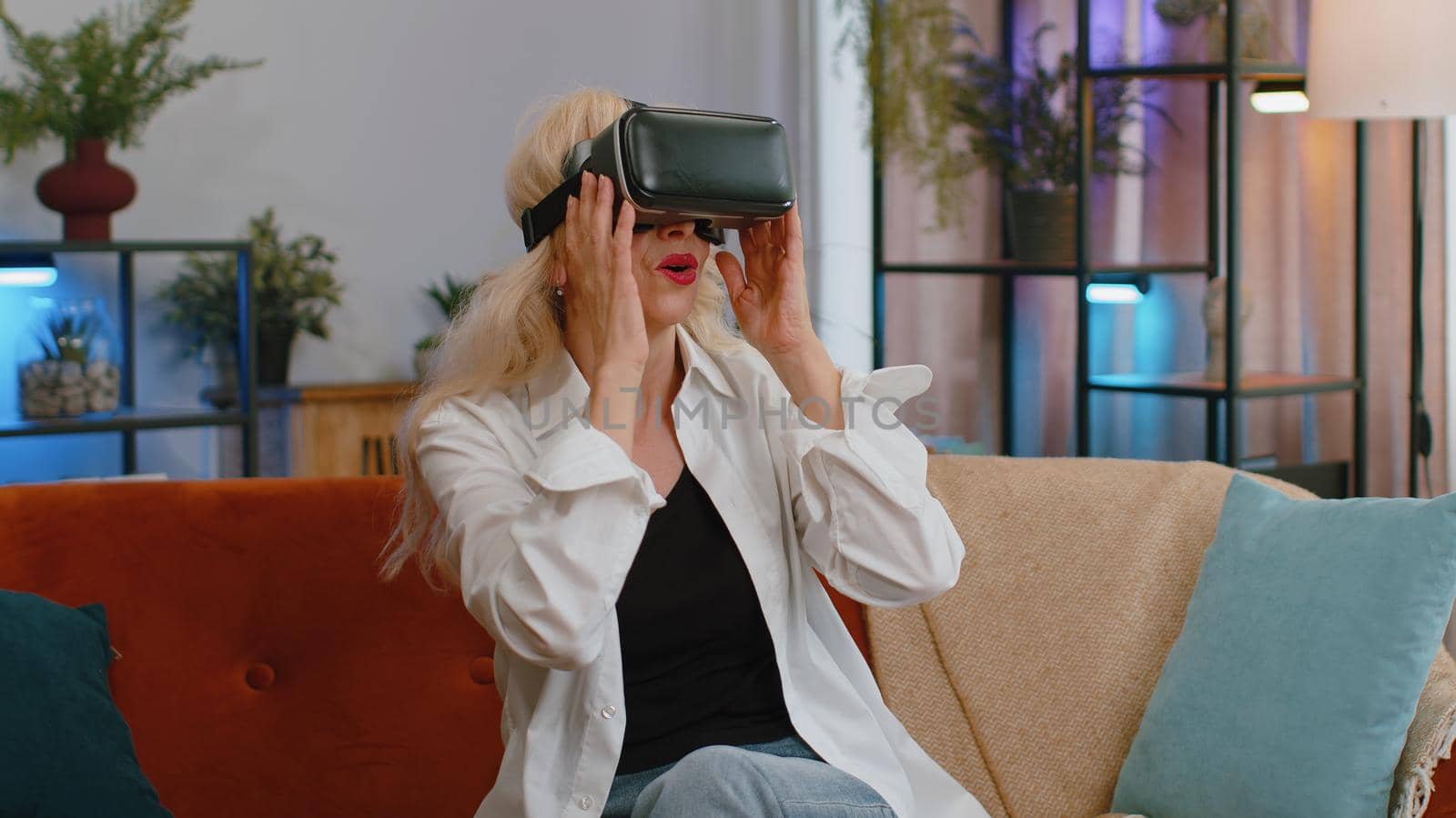 Senior old grandmother using virtual reality futuristic technology VR app headset helmet to play simulation 3D video game watching film movie at modern home apartment. Woman in goggles sitting on sofa