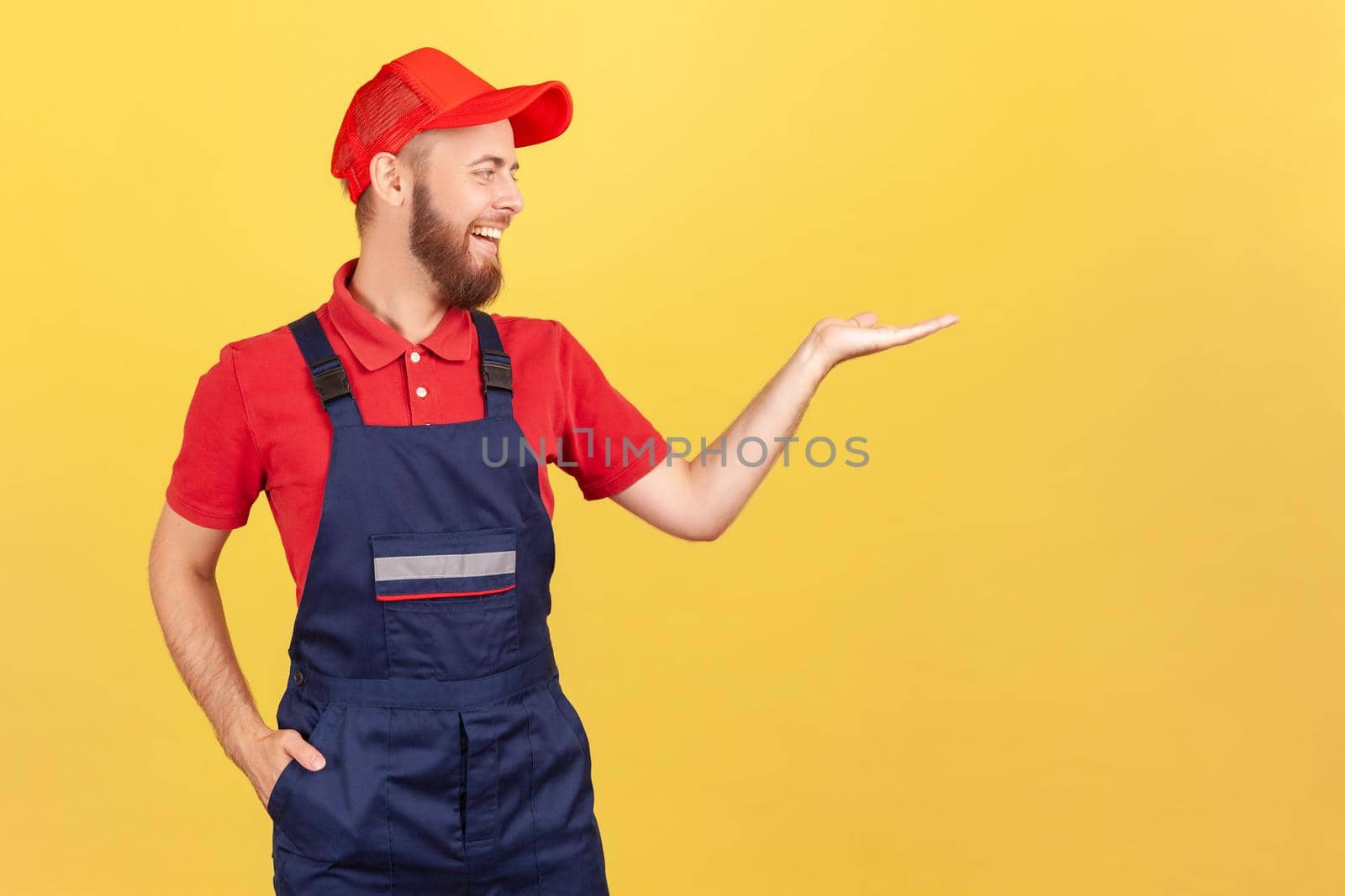 Portrait of smiling worker man standing and presenting copy space for advertisement on his palm, looking away, wearing overalls and red cap Indoor studio shot isolated on yellow background.