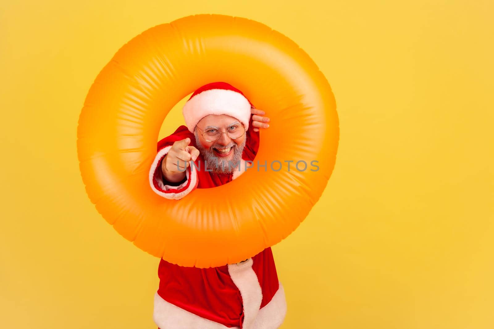 Happy elderly man with gray beard in santa claus costume standing with rubber ring, pointing to camera with toothy smile, having positive expression. Indoor studio shot isolated on yellow background.