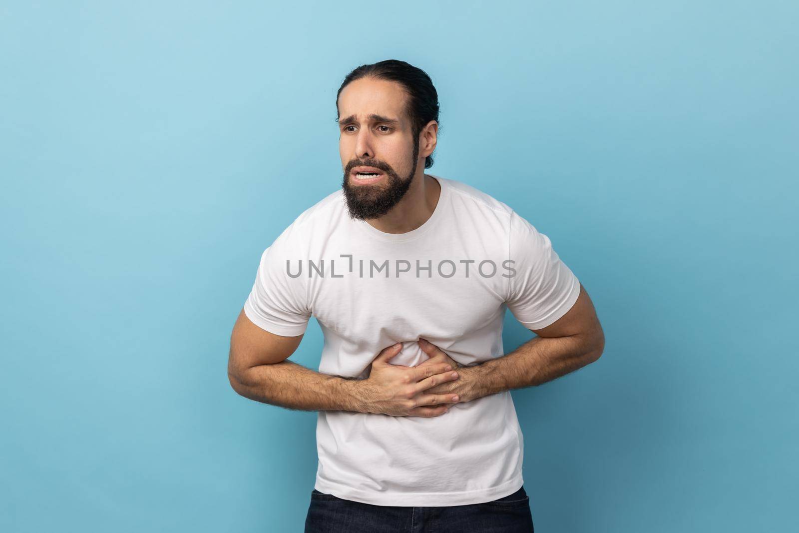 Portrait of ill sick man with beard wearing white T-shirt grimacing and suffering from pain in belly, severe abdominal distress Indoor studio shot isolated on blue background.