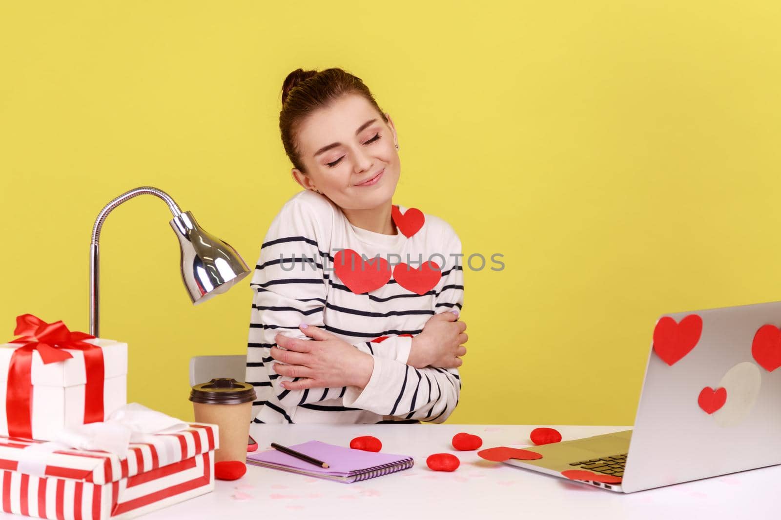 Woman covered with heart sticks sitting at workplace, hugging herself and smiling, feeling comfortable and fulfilled, egoistic person. Indoor studio studio shot isolated on yellow background.