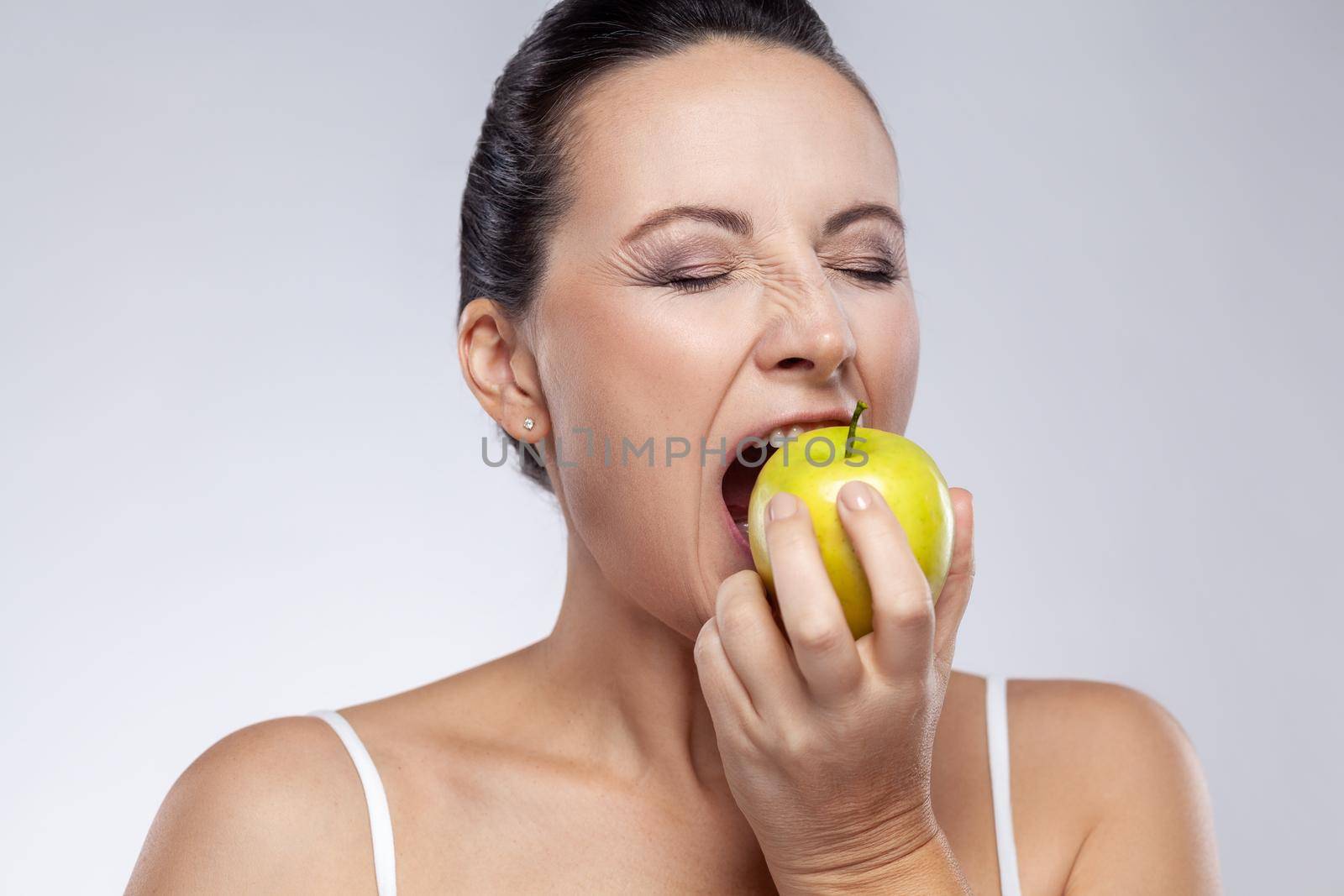 Closeup portrait of middle aged woman biting fresh green apple, uses natural cosmetic, healthy eating, vitamins for health and skin care. Indoor studio shot isolated on gray background.