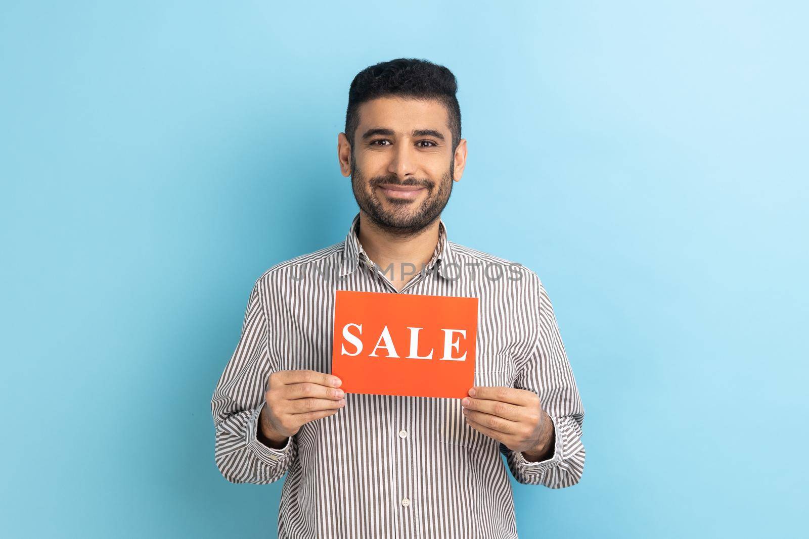 Smiling businessman holding card with Sale word, feeling carefree amused by discounts, Black Friday low prices, wearing striped shirt. Indoor studio shot isolated on blue background.