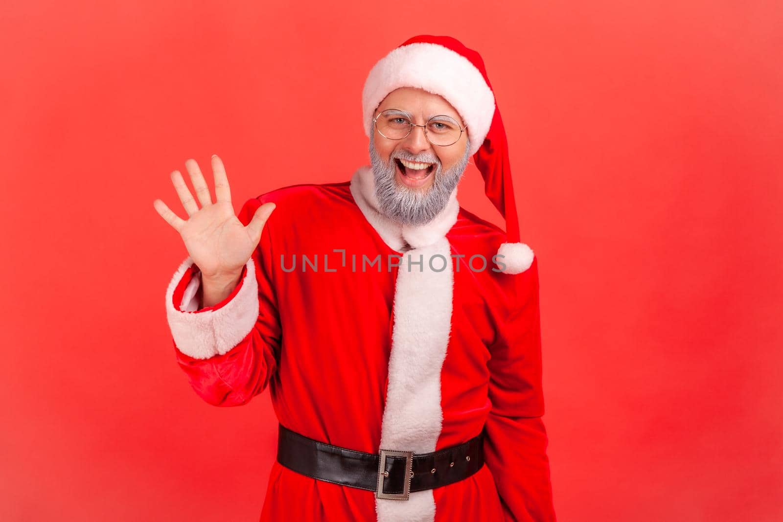 Hello. Happy elderly man with gray beard wearing santa claus costume raising palm to wave hi, greeting with hospitable friendly smile. Indoor studio shot isolated on red background.