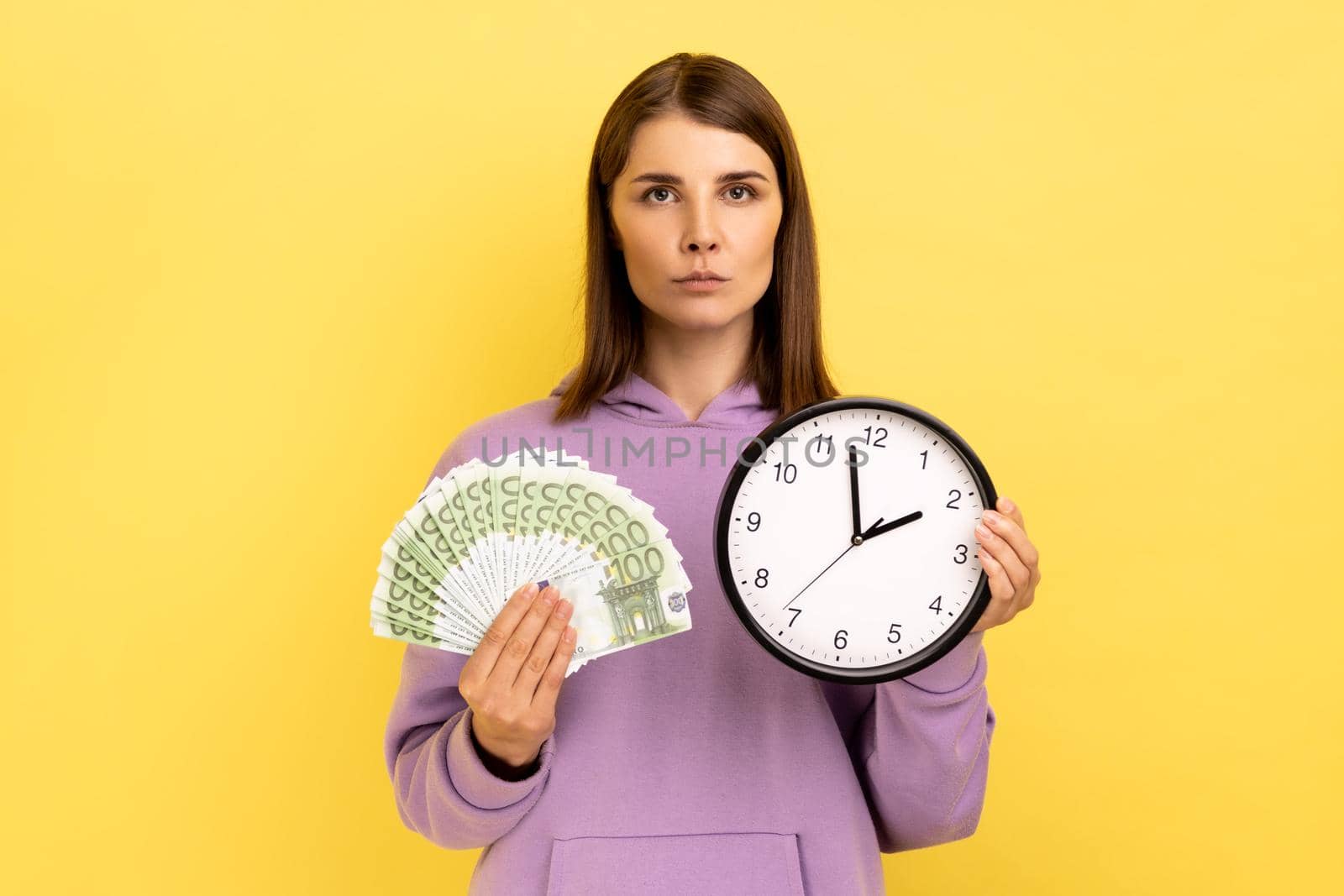 Portrait of rich dark haired woman holding big fan of euro banknotes and wall clock, time is money, looking seriously at camera, wearing purple hoodie. Indoor studio shot isolated on yellow background