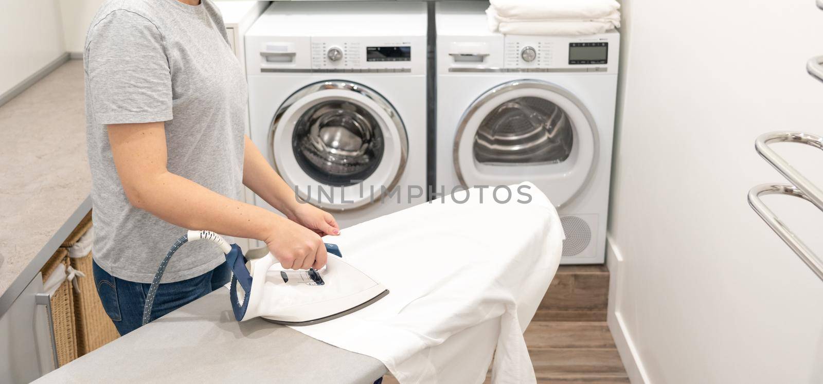 Woman ironing white clothes on board in laundry room with washing machine on background by Mariakray