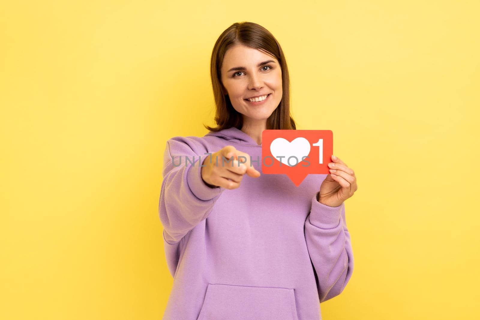 Portrait of dark haired positive woman blogger holding hear like icon and pointing finger at camera with toothy smile, wearing purple hoodie. Indoor studio shot isolated on yellow background.