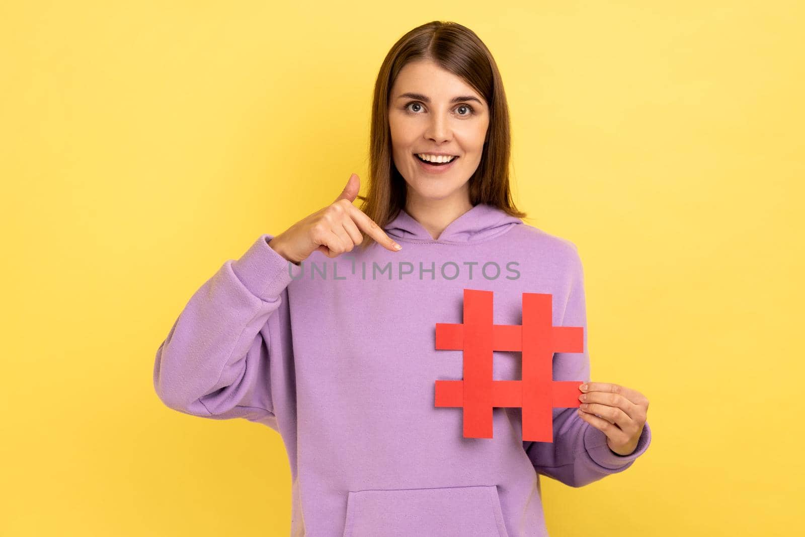 Attention to interesting blog post. Happy woman pointing at red hashtag symbol, making important topic popular, setting trends on Internet. Indoor studio shot isolated on yellow background.