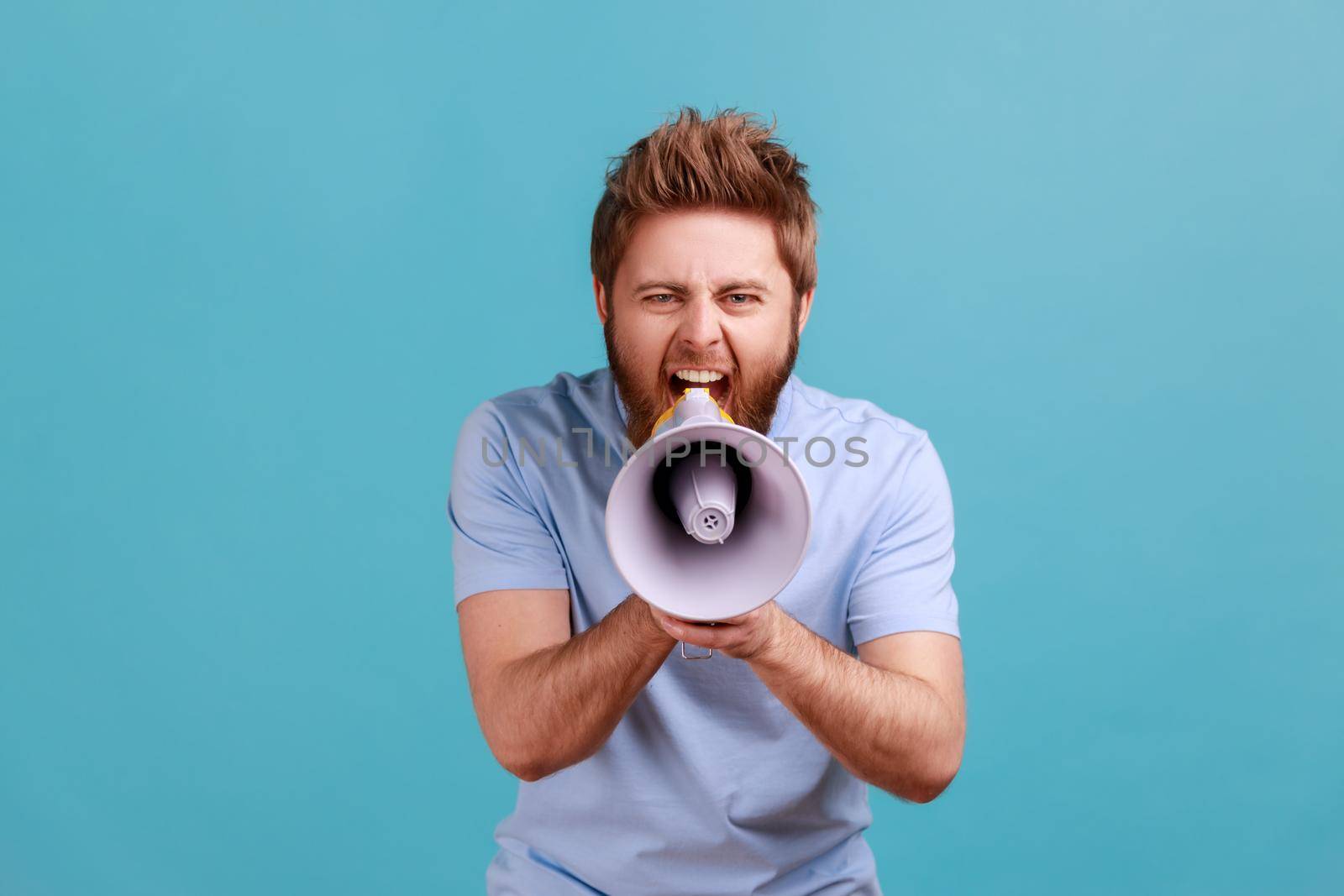 Portrait of bearded man holding megaphone near mouth, loudly speaking, screaming, making announcement, paying attention at social problems. Indoor studio shot isolated on blue background.