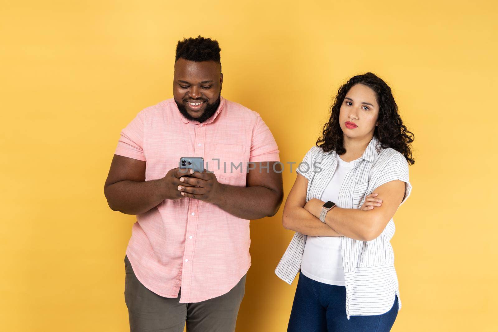Satisfied young couple in casual clothing standing together, man using mobile phone and smiling, offended lonely woman standing with folded arms. Indoor studio shot isolated on yellow background.