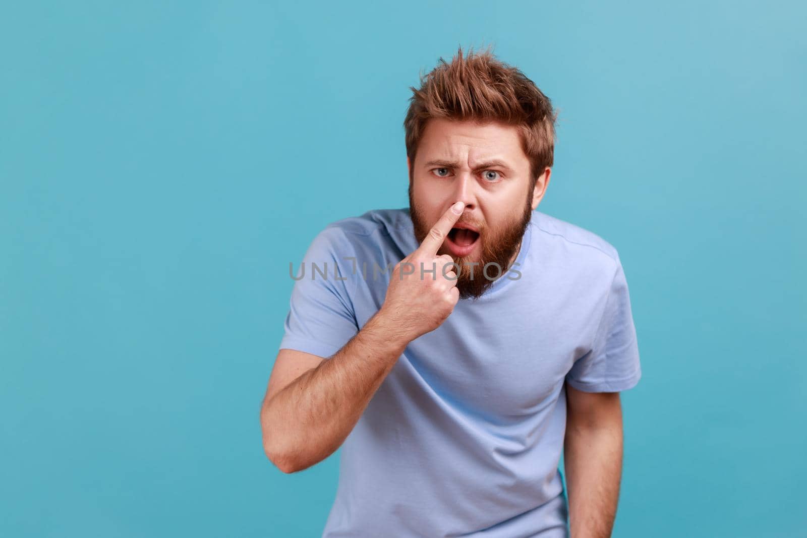 You are liar. Portrait of shocked bearded man standing, touching his nose and showing lie gesture, suspecting falsehood, dishonest talk. Indoor studio shot isolated on blue background.